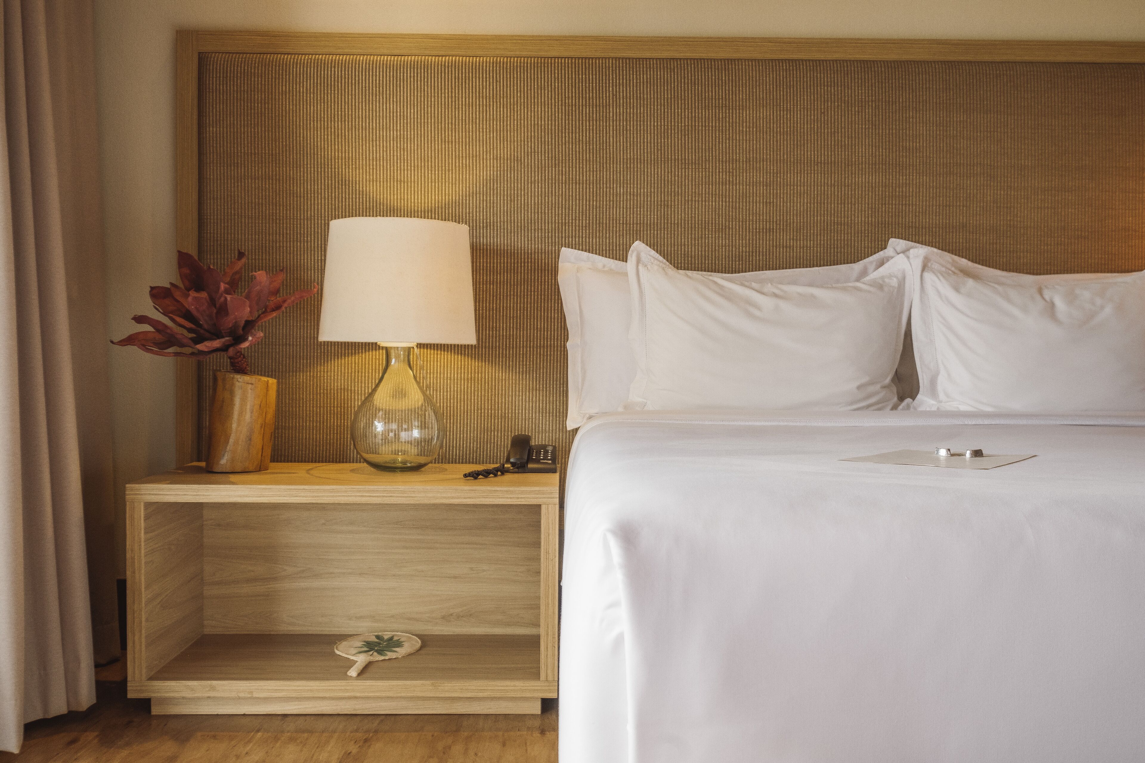 A hotel room with a bed, a nightstand with a lamp, a vase with dried flowers, and a remote control.