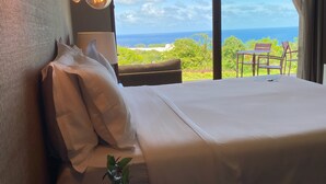 A bedroom with a large bed, a small round table with a vase, and a view of the ocean through a large window. - Colina Pousada SPA (Fernando de Noronha)