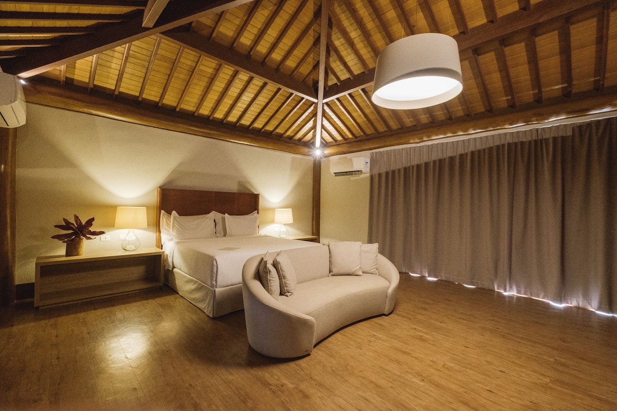 Une chambre à coucher comprenant un lit, un canapé, une table de chevet avec une lampe et une grande fenêtre avec des rideaux.