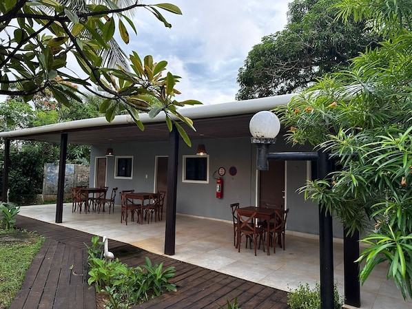 A covered outdoor dining area with wooden tables and chairs, surrounded by lush greenery and a stone wall.