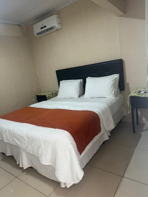 A single bed with white bedding and a brown accent, a nightstand with a lamp, and a wall-mounted air conditioner.