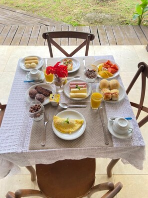 A table set with breakfast items including eggs, pastries, and fruit, on a patio with chairs and a view of the outdoors. - Pousada da Jo (Fernando de Noronha)