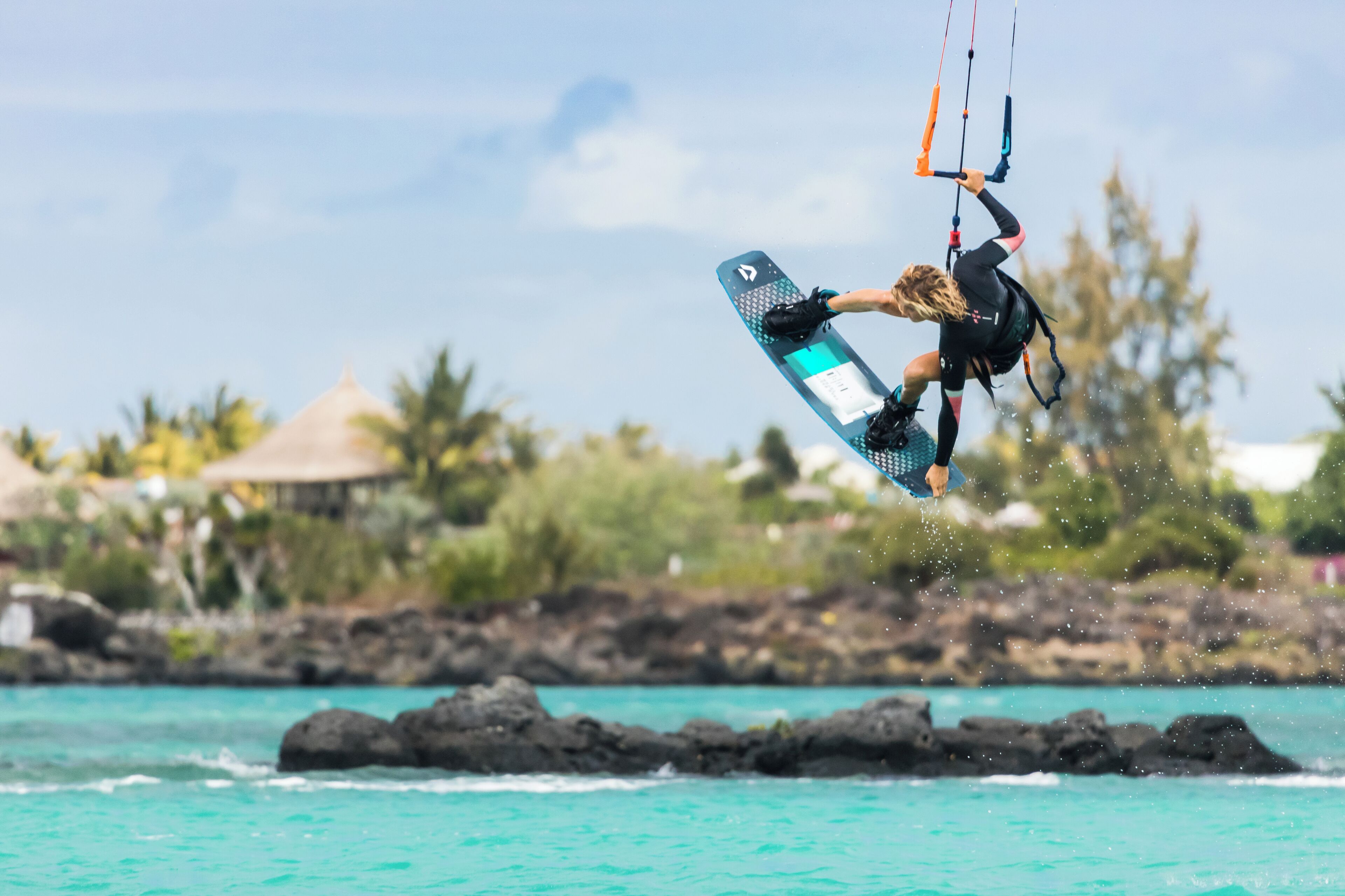 En person kitesurfer over turkis vand med en klippefyldt kystlinje og hytter med stråtag i baggrunden.