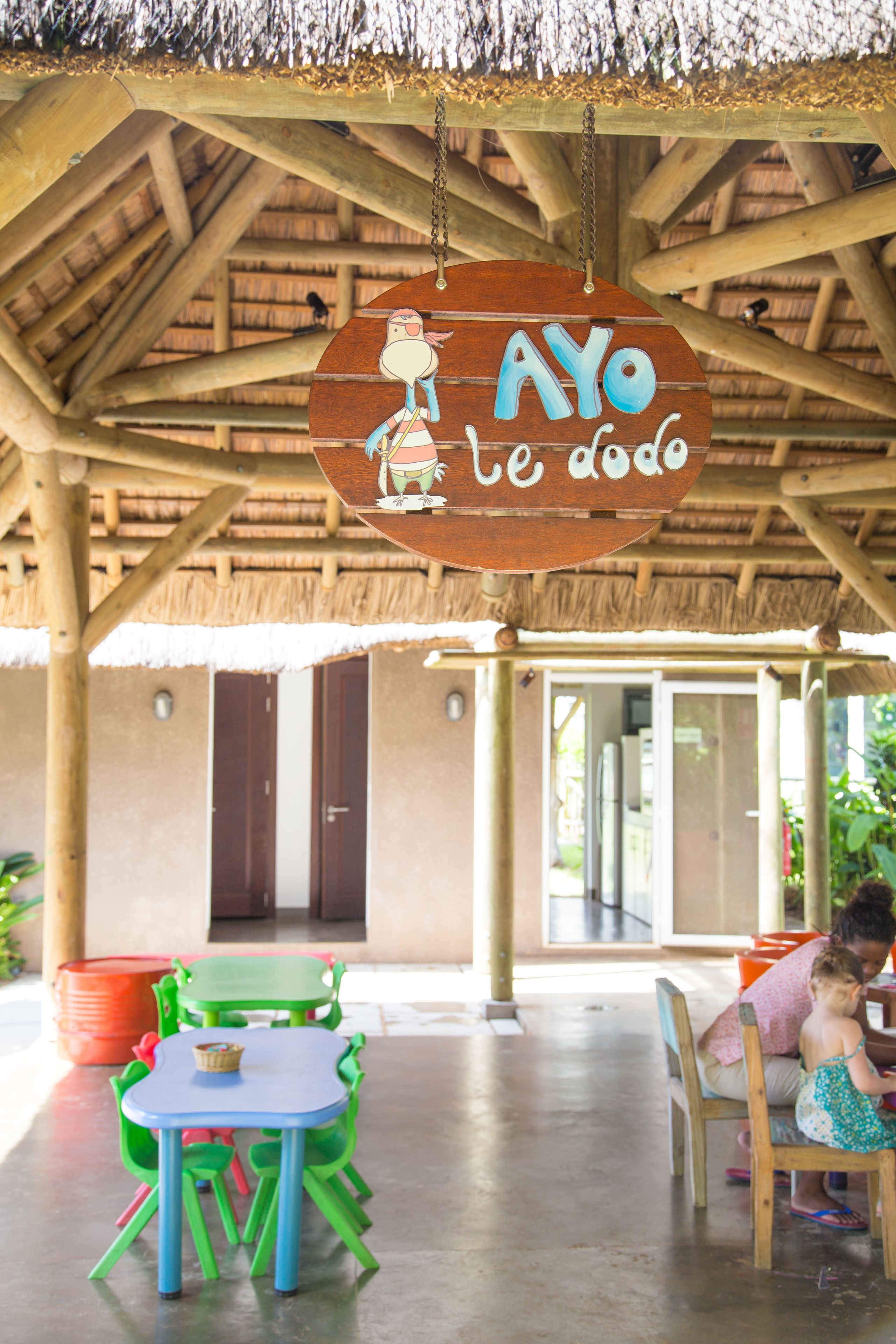 Une structure en bois surmontée d’un toit de chaume, une enseigne indiquant « AYO te doo », et une table avec des chaises.