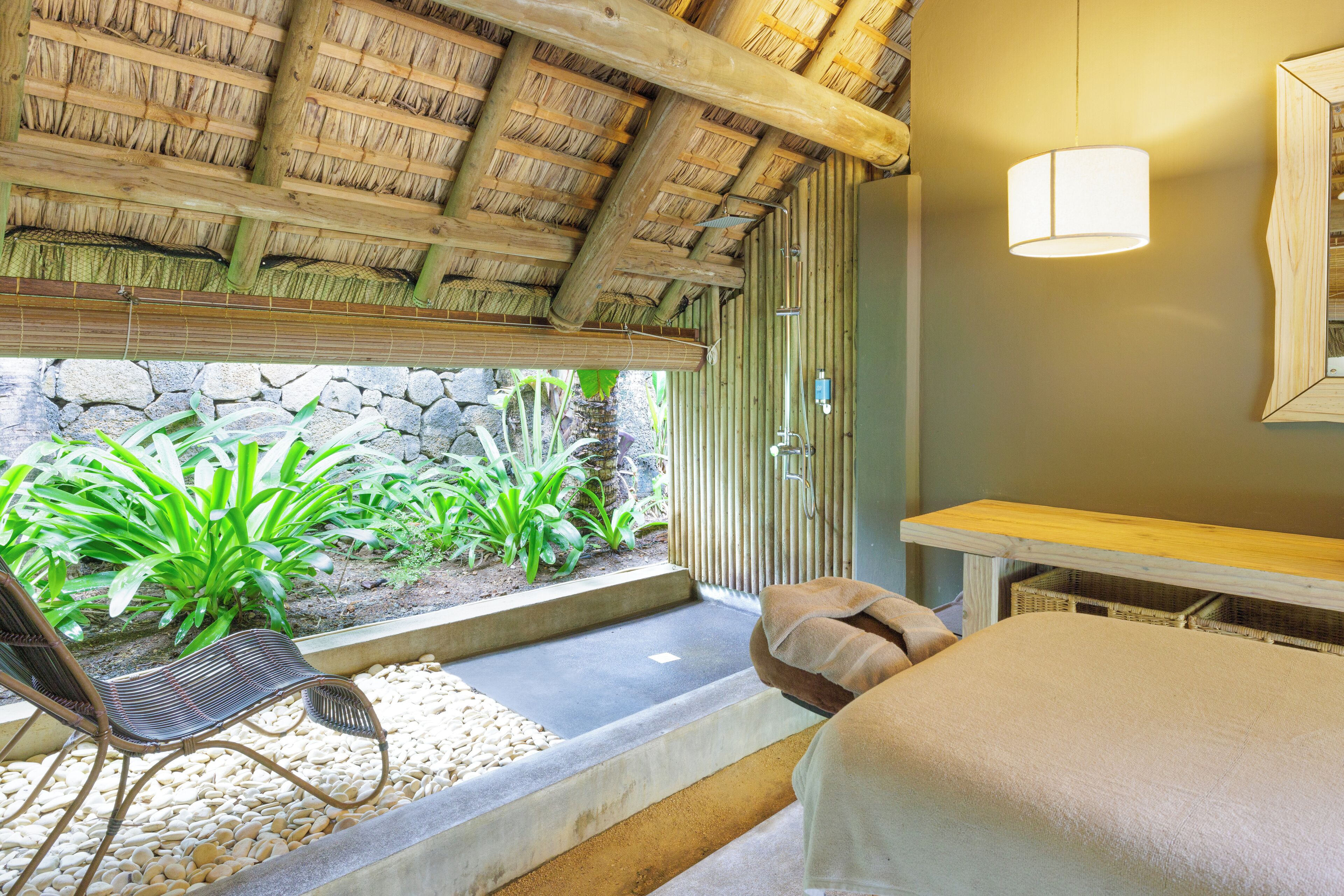 A room with a bed, a chair, a table, a stone wall, and a thatched roof.