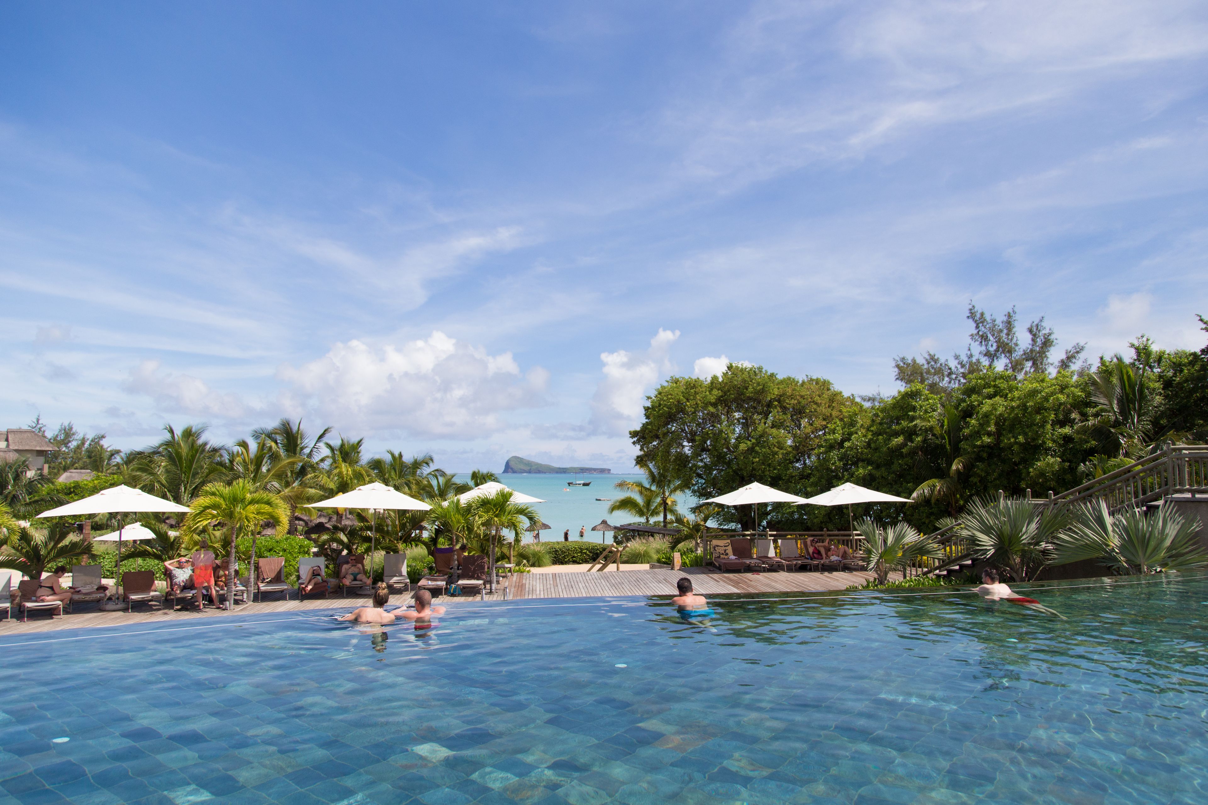 A clear blue pool with lounge chairs and umbrellas, surrounded by palm trees and a view of the ocean.