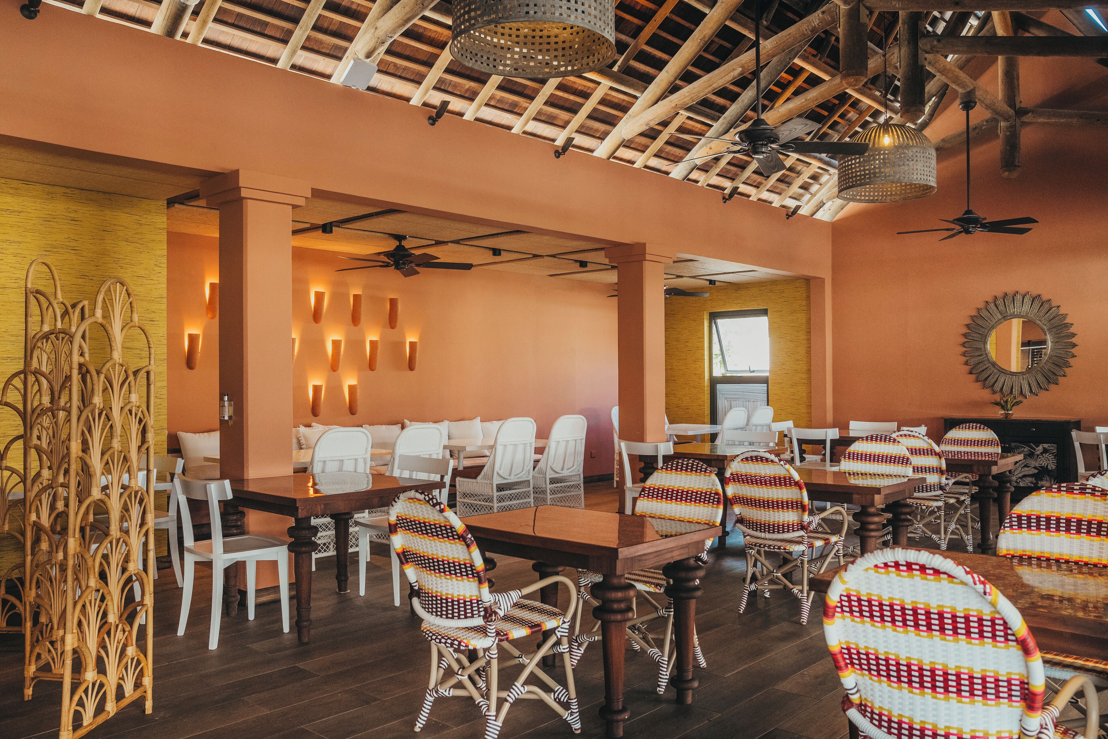 Un restaurante con mesas de madera y sillas de mimbre, un ventilador de techo e iluminación en las paredes.