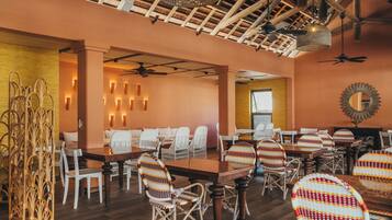 Un restaurante con mesas de madera y sillas de mimbre, un ventilador de techo e iluminación en las paredes.