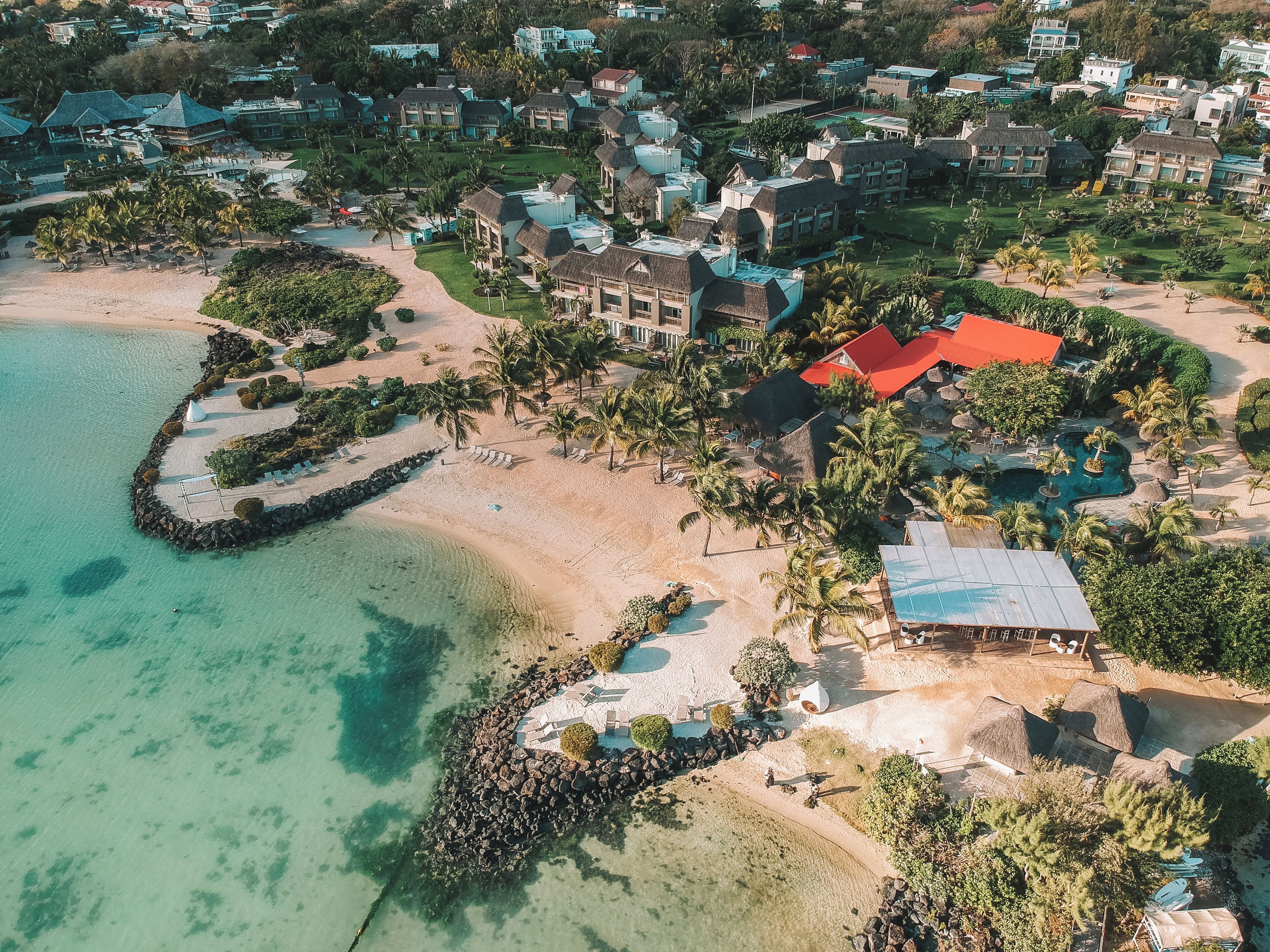 Vista aérea de un complejo turístico costero con propiedades frente a la playa, un rompeolas rocoso y un mar azul cristalino.