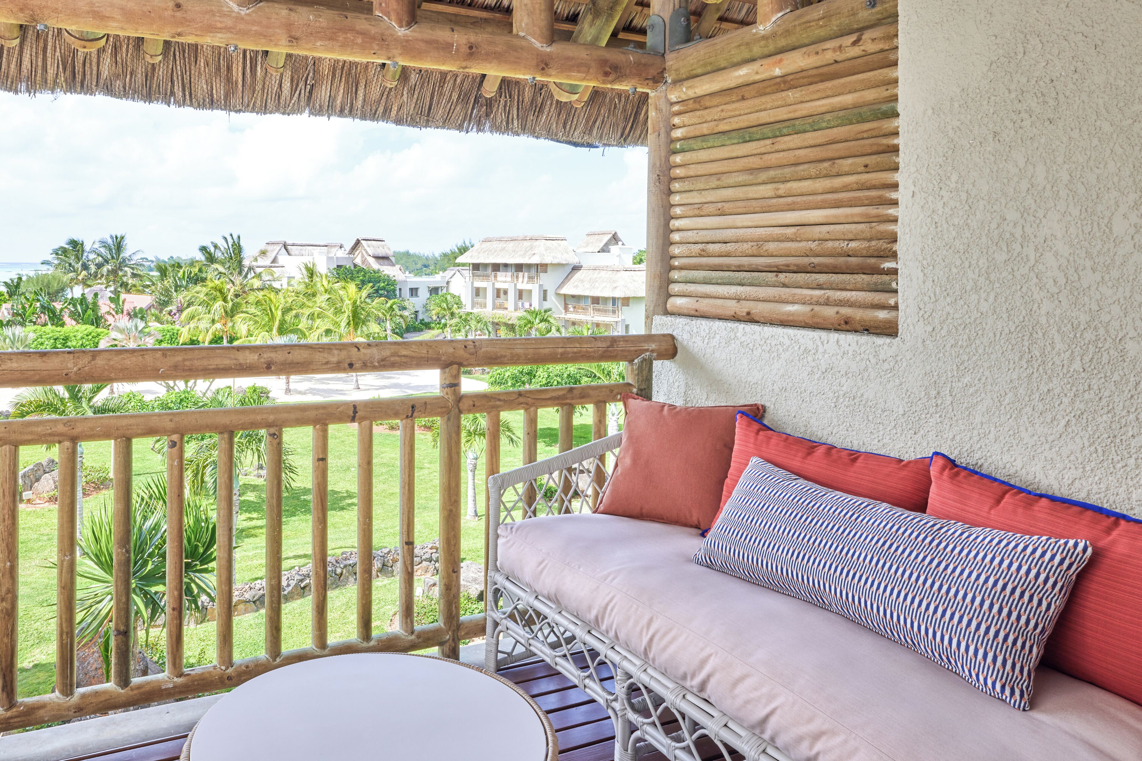 A balcony with a wicker sofa, a round table, and a view of a resort with palm trees and buildings.
