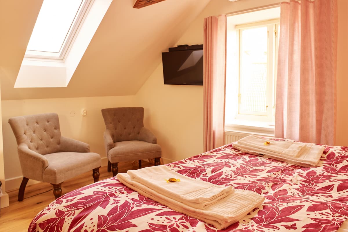 A bedroom with a sloped ceiling, two armchairs, a bed with a floral bedspread, and a window with curtains.