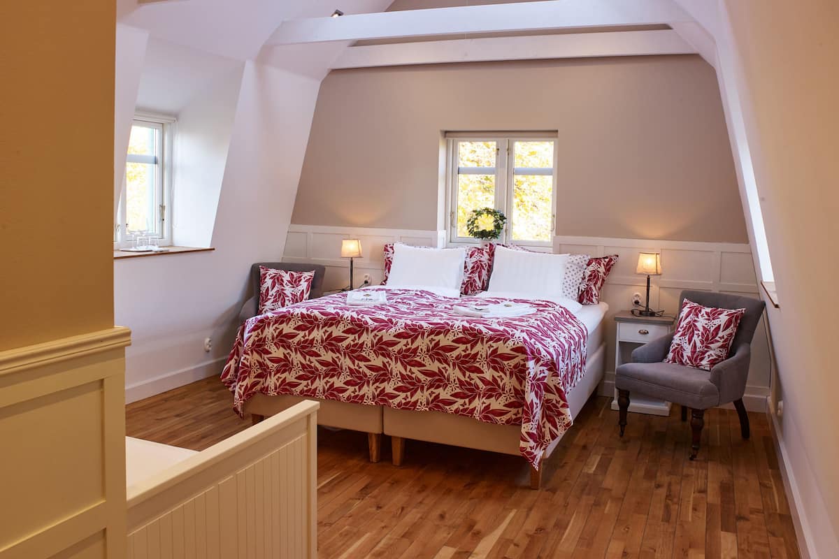 A bedroom with a sloped ceiling, a bed with a floral bedspread, two bedside tables with lamps, a chair, and a window with a wreath.