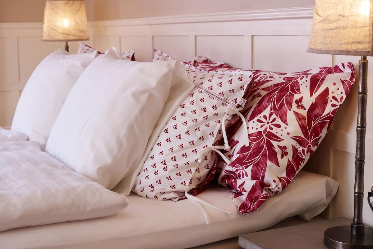 A neatly made bed with white and red patterned pillows, a bedside table with a lamp, and a wall with a decorative panel.
