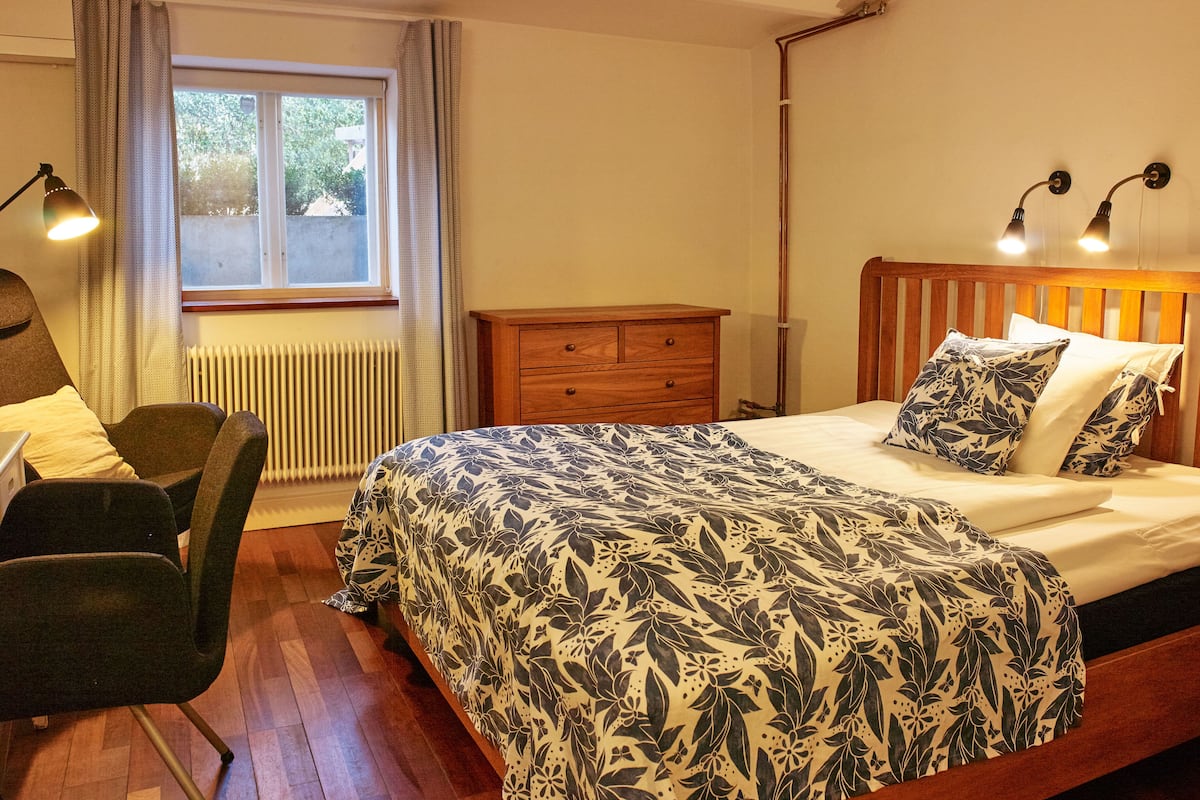 A bedroom with a wooden bed, a nightstand, a chair, a window with curtains, and a radiator.