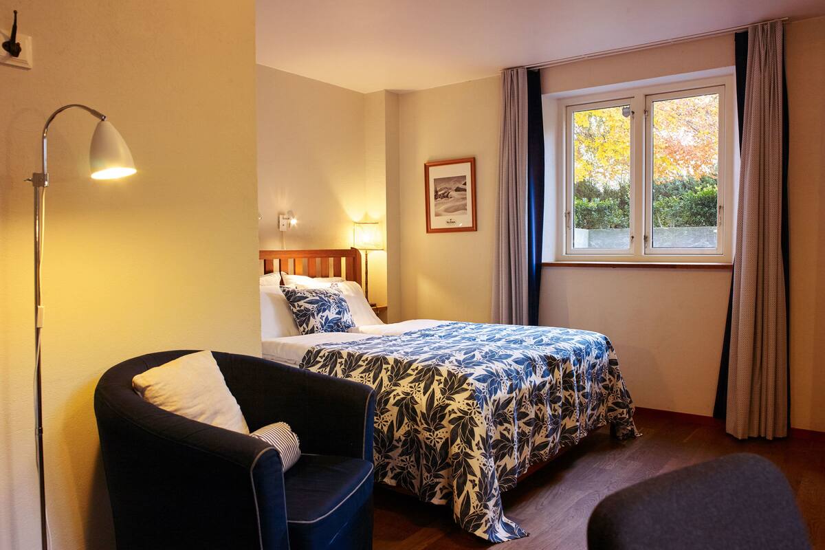 A hotel room with a bed, a chair, a lamp, a window with curtains, and a framed picture on the wall.