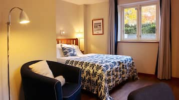 Une chambre d’hôtel avec un lit, une chaise, une lampe, une fenêtre avec des rideaux et un tableau encadré au mur.