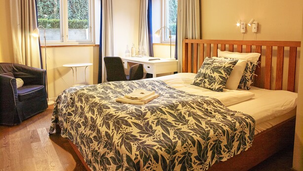A hotel room with a bed, a desk, a chair, and a view of greenery outside. - MillasVilla (Lund)