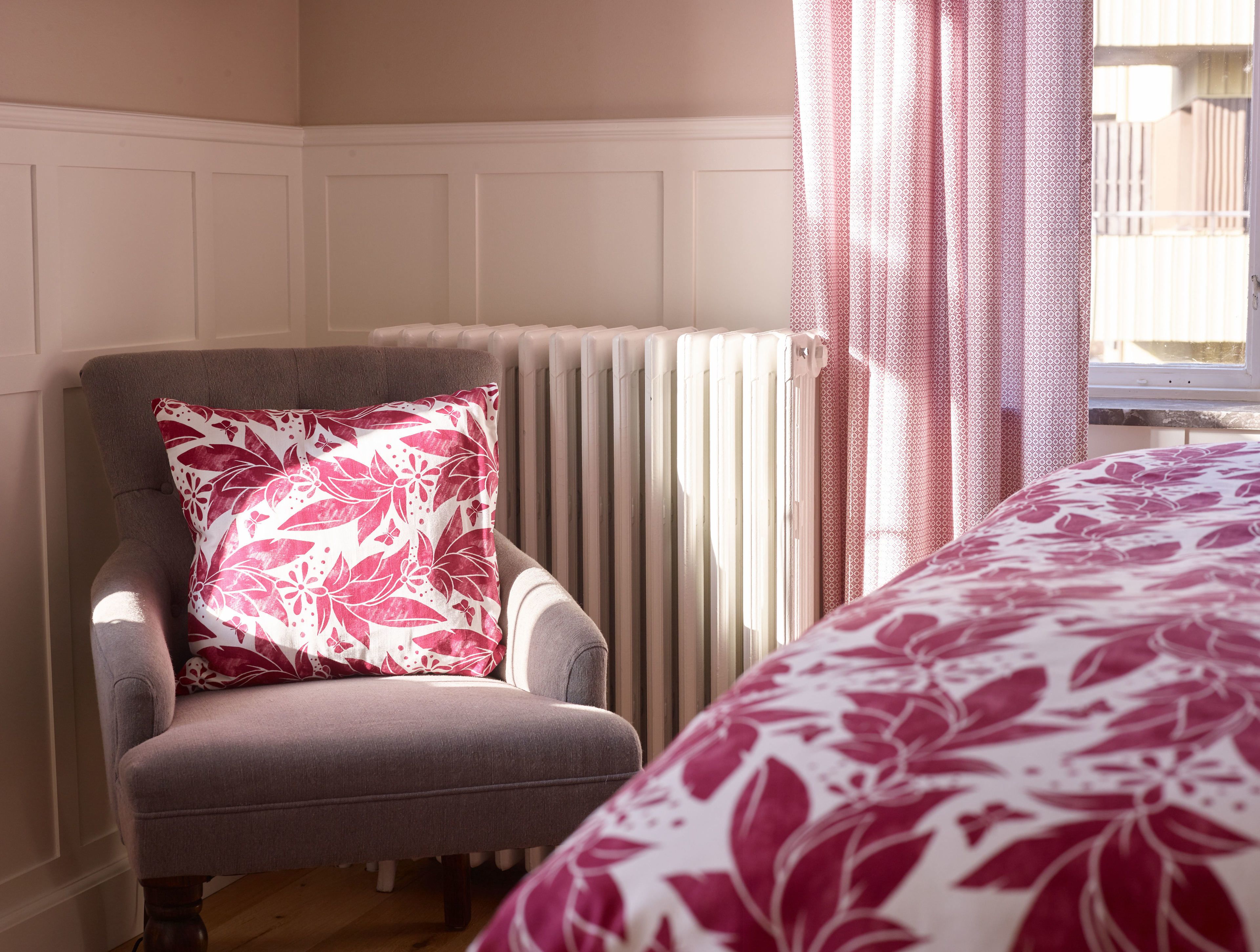 Une pièce avec un fauteuil gris, un radiateur et un lit recouvert d’une couvre-lit à motifs floraux.