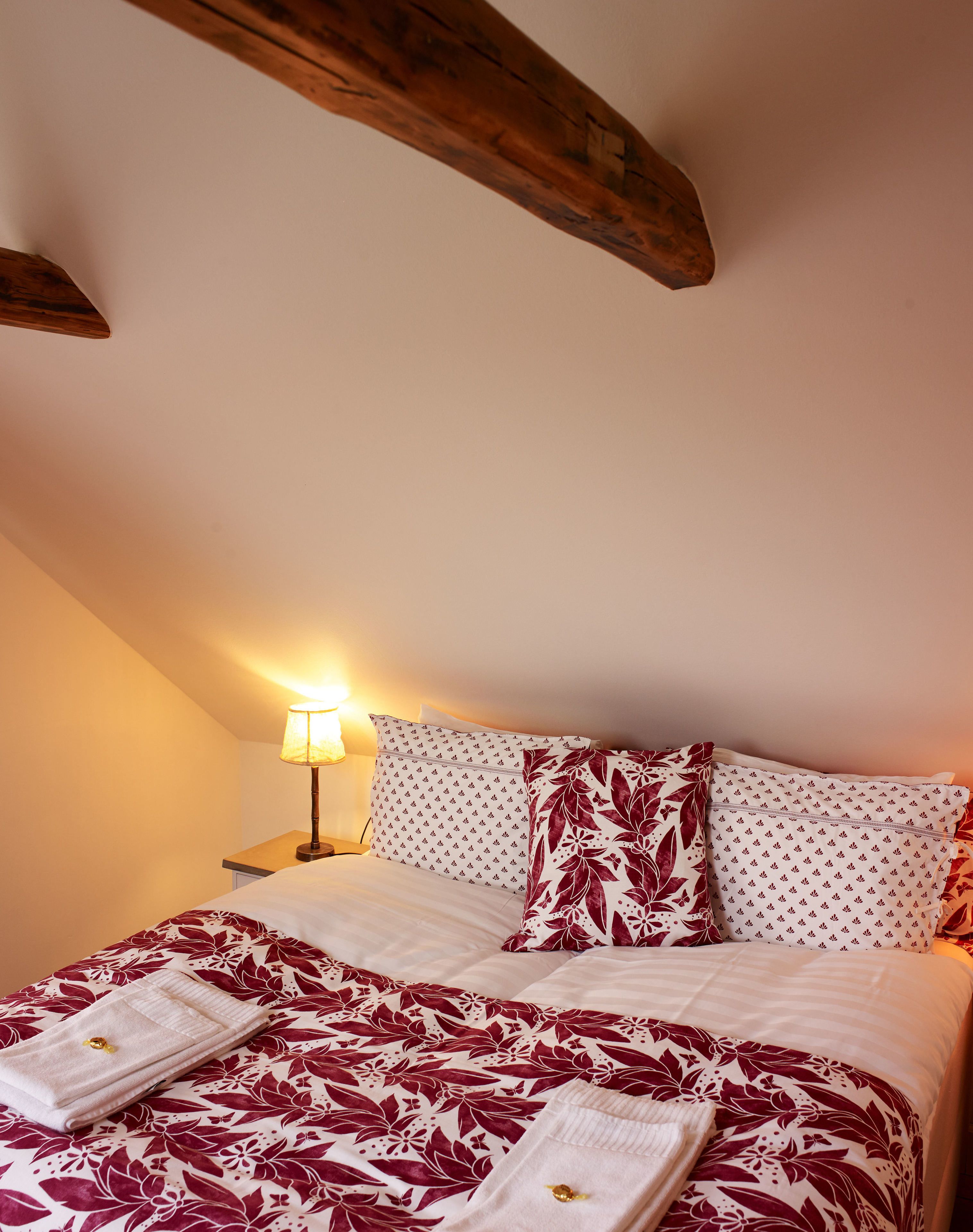 Una cama bien hecha con ropa de cama floral, una viga de techo de madera y una lámpara de noche.