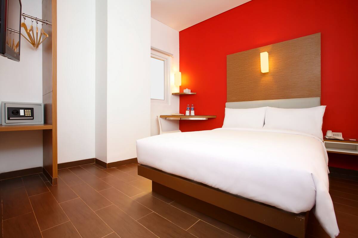 Habitación de hotel con una cama grande, una mesita de noche con lámpara, un estante con botellas y un panel de control en la pared.