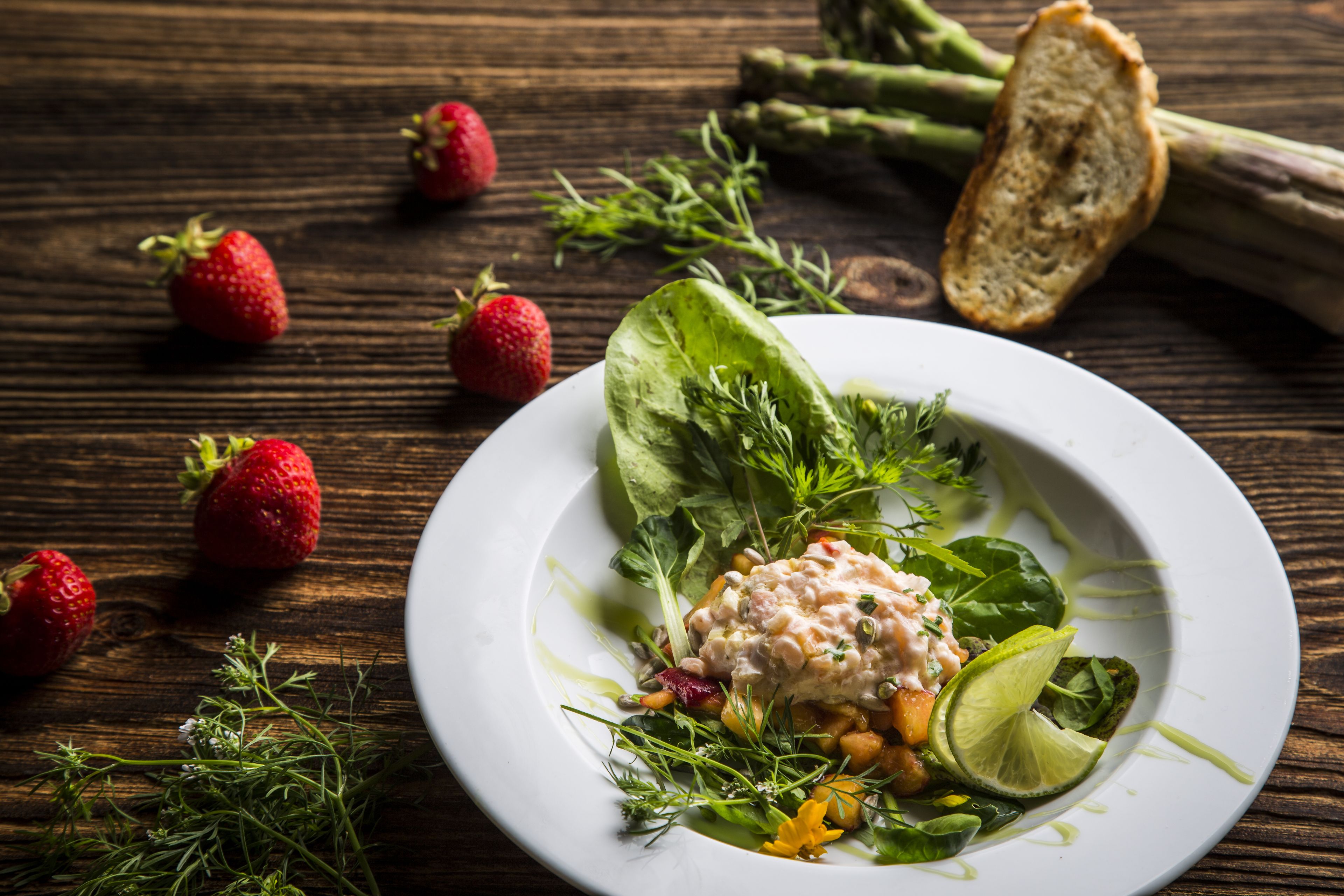 Une assiette de nourriture contenant des fraises, des asperges et un quartier de citron vert, posée sur une table en bois.