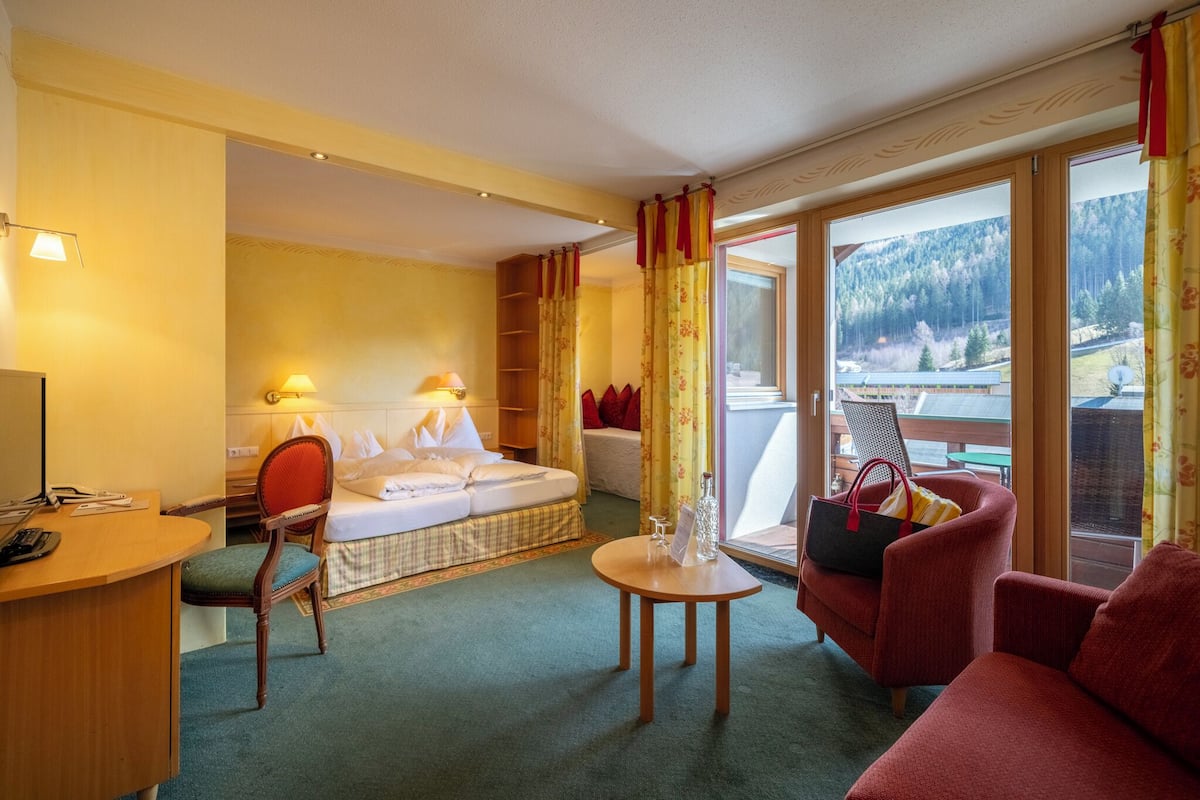 A hotel room with a bed, a desk, a chair, a sofa, and a balcony with a view of the mountains.