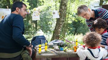 Un grupo de personas reunidas alrededor de una mesa con diversos objetos, como botellas y flores, en un entorno natural con árboles al fondo.