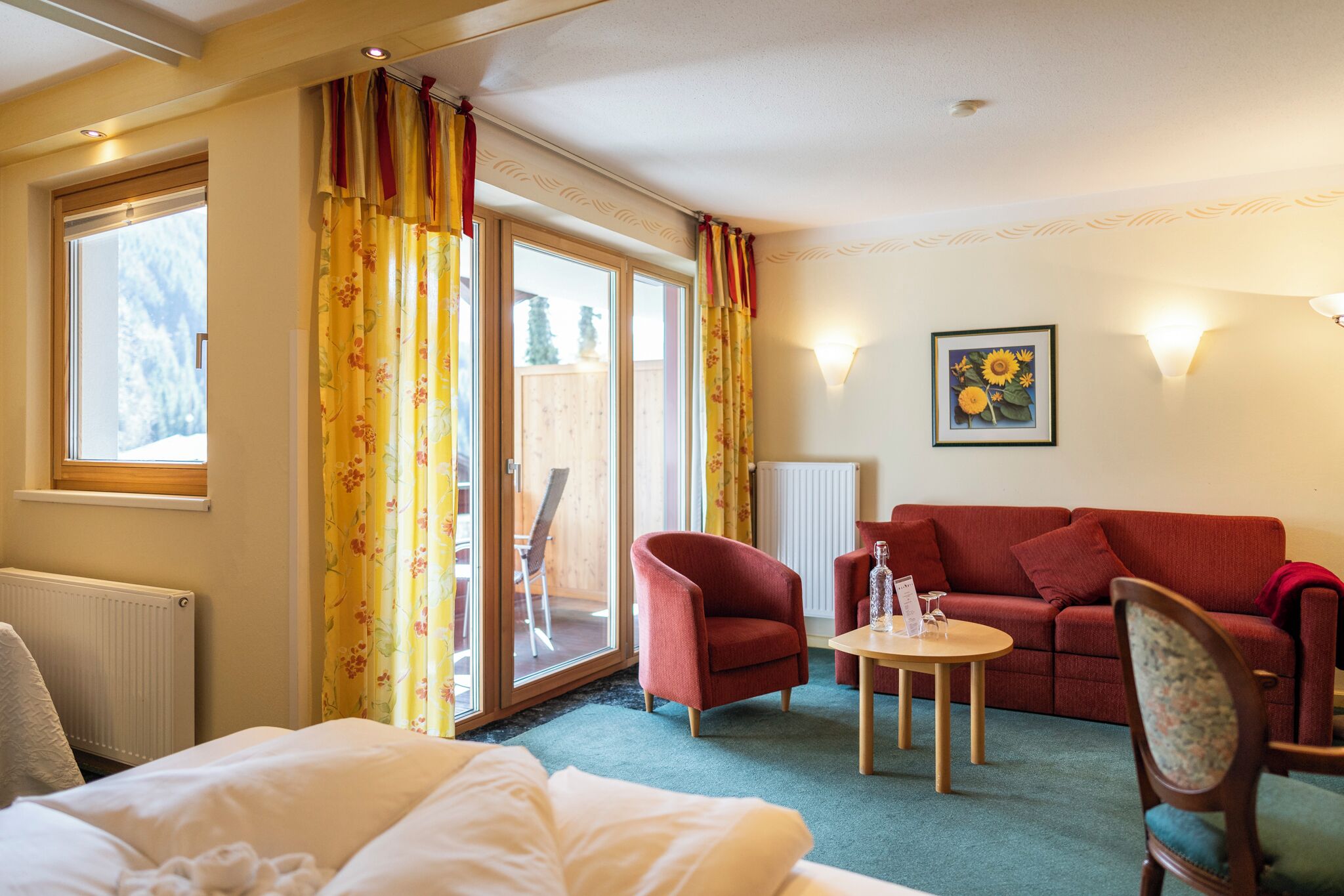 Et hotelværelse med en seng, en rød sofa, et lille bord og en stol. Der er et vindue med gardiner, en radiator og en vægmonteret lampe.