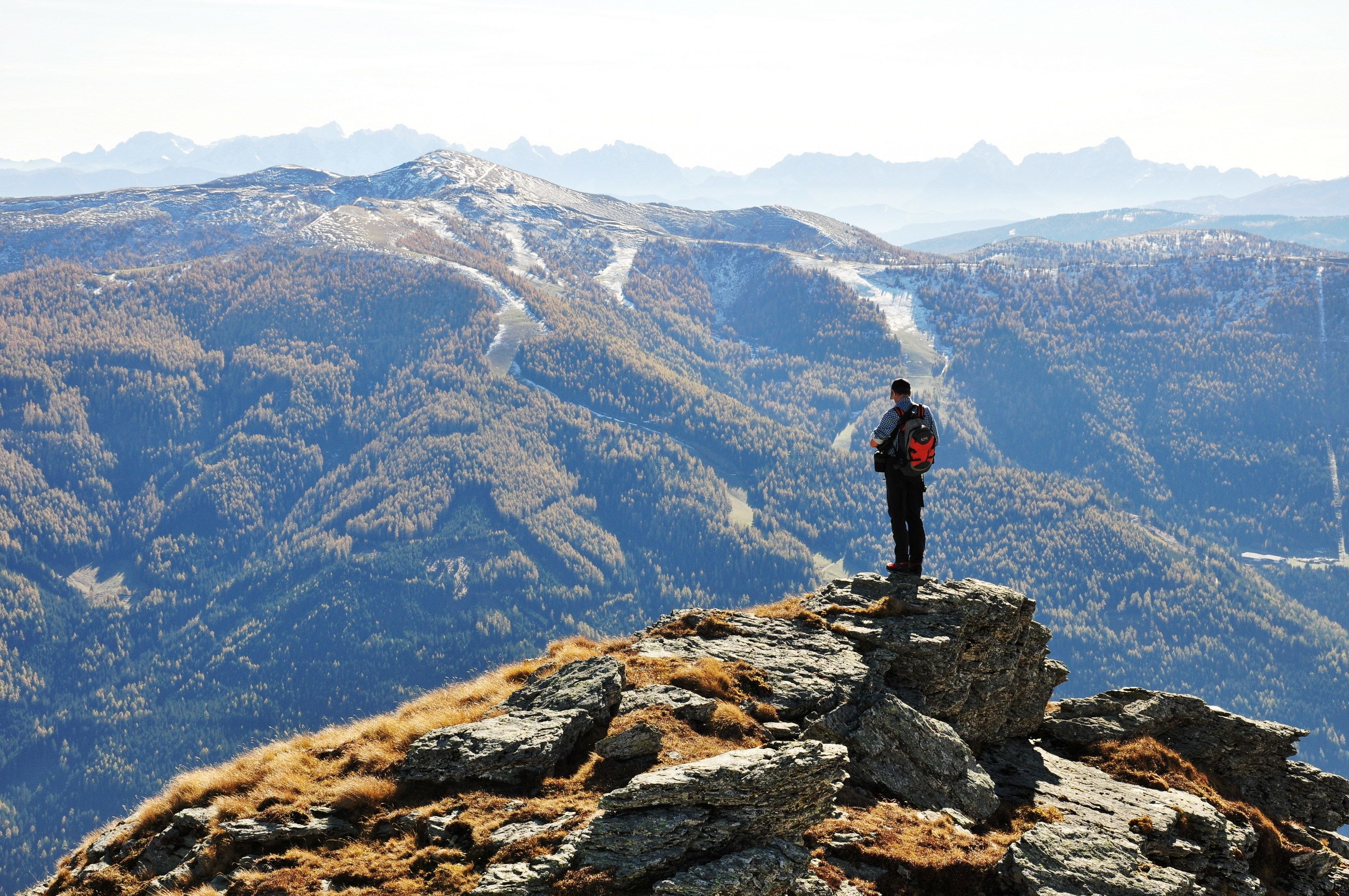 Une personne se tient sur un promontoire rocheux, un sac à dos sur le dos, et surplombe un paysage montagneux avec une vallée en contrebas.