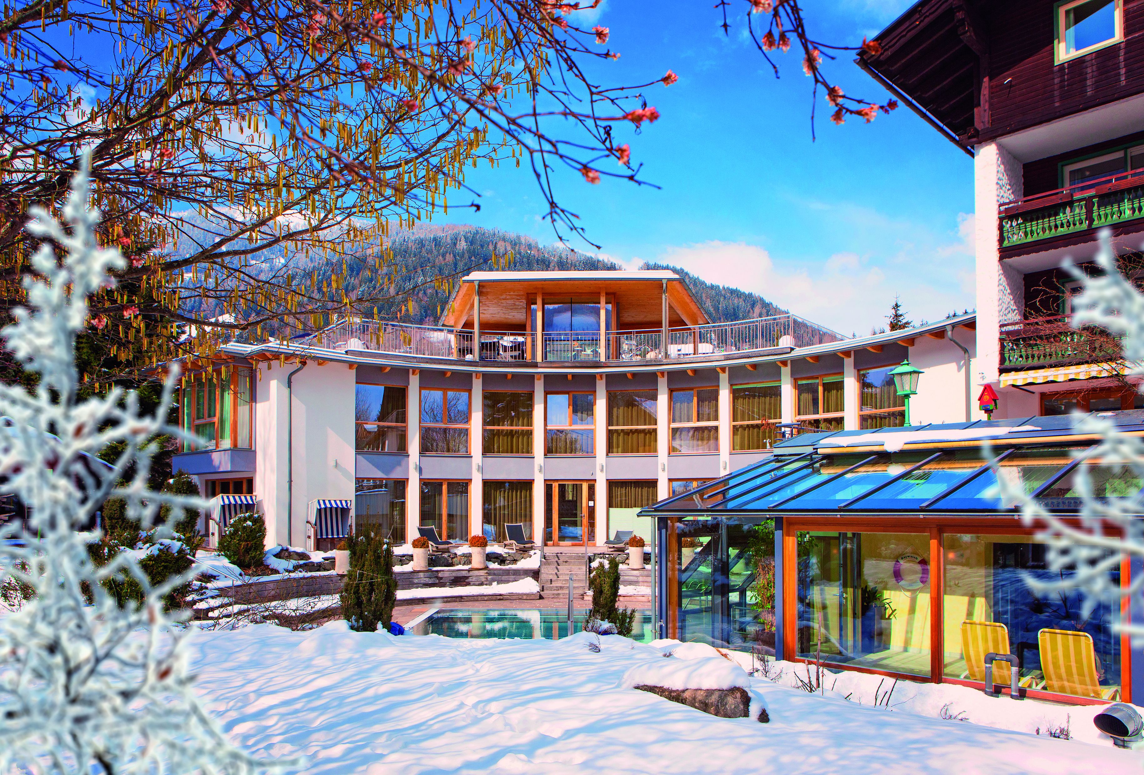 Een modern hotel met een glazen zwembad, meerdere balkons en uitzicht op met sneeuw bedekte bergen.
