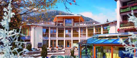 A modern hotel with a glass-enclosed pool, multiple balconies, and a view of snow-covered mountains.
