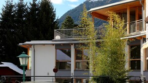 Una terraza de madera con sillones de descanso, un edificio con balcón y una montaña nevada al fondo.