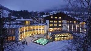 Un hotel moderno con fachada de vidrio, piscina exterior climatizada y montañas al fondo.