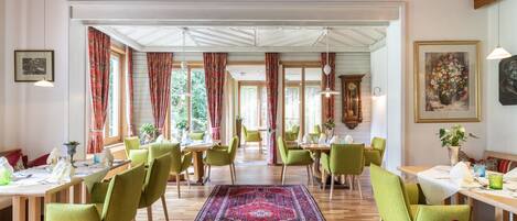 A dining area with wooden tables and green chairs, a patterned rug, and large windows with curtains.
