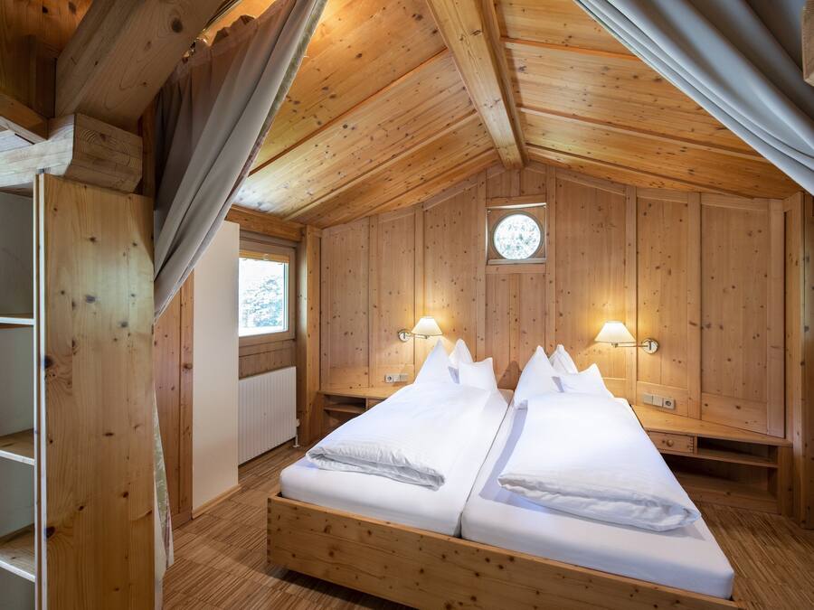 Ein Schlafzimmer in einer Holzhütte mit einem Bett, zwei Nachttischlampen, einem Fenster und einem runden Dachfenster.