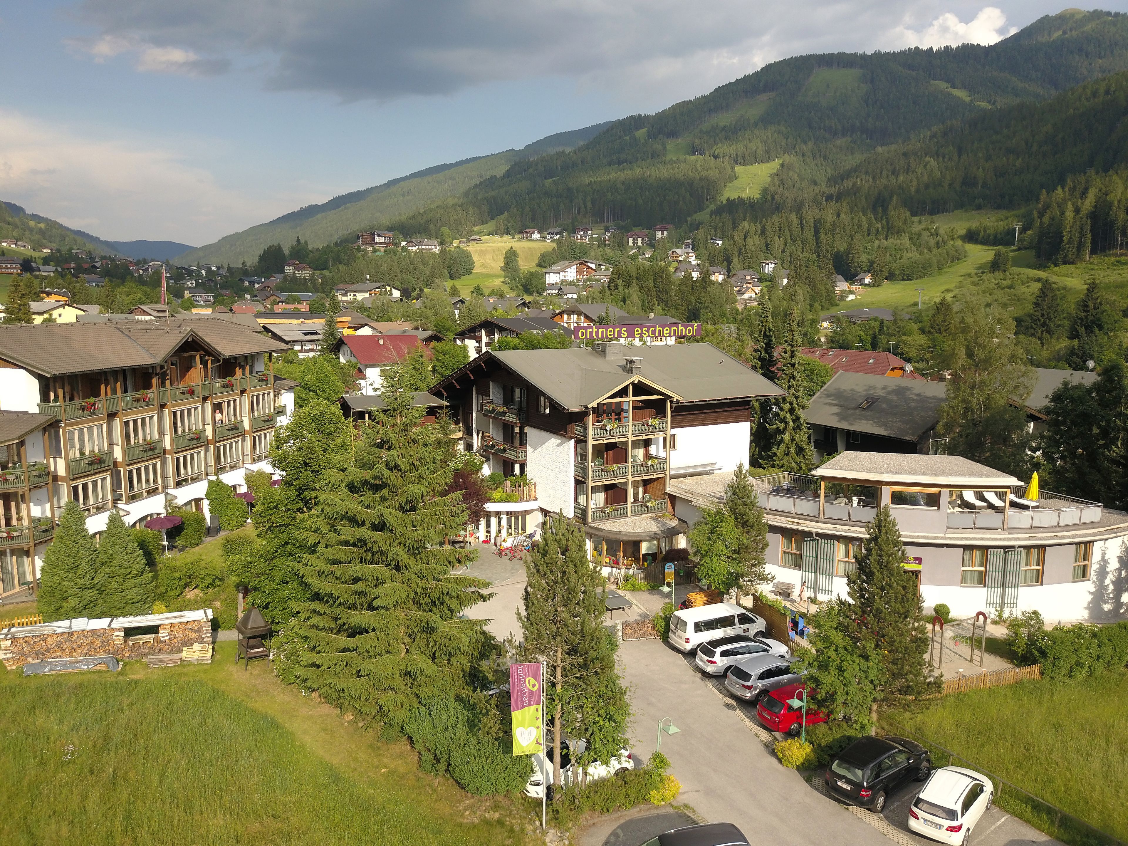 A mountain resort with chalet-style buildings, a parking area, and a view of the surrounding forest and hills.