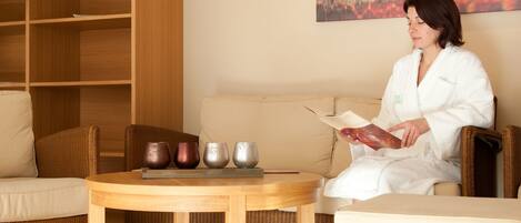 A person in a white robe sitting on a beige sofa in a well-lit room with a wooden bookshelf, a round wooden table with teapots, and a large abstract painting on the wall.