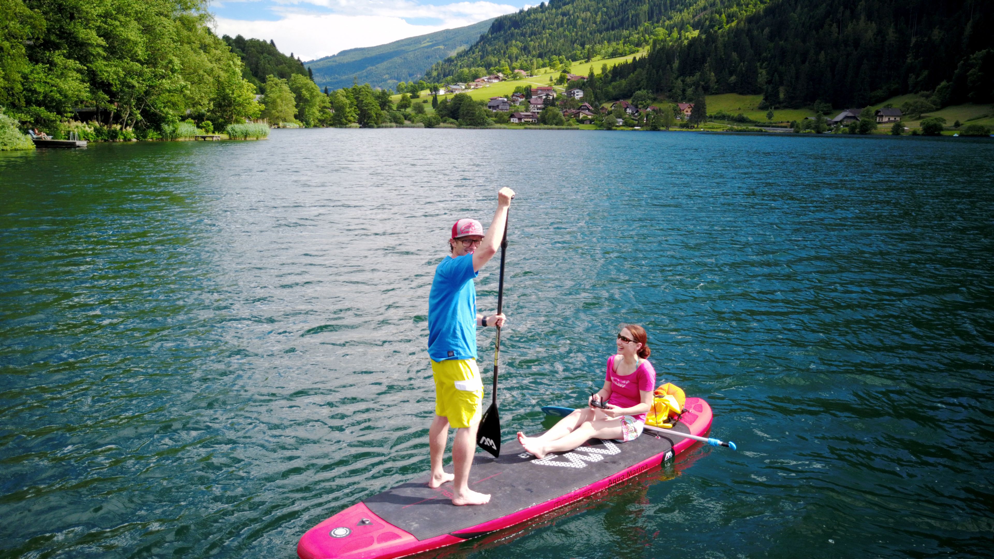 Ein Paar steht auf einem Stand-Up-Paddleboard auf einem ruhigen See, im Hintergrund sind Häuser zu sehen.