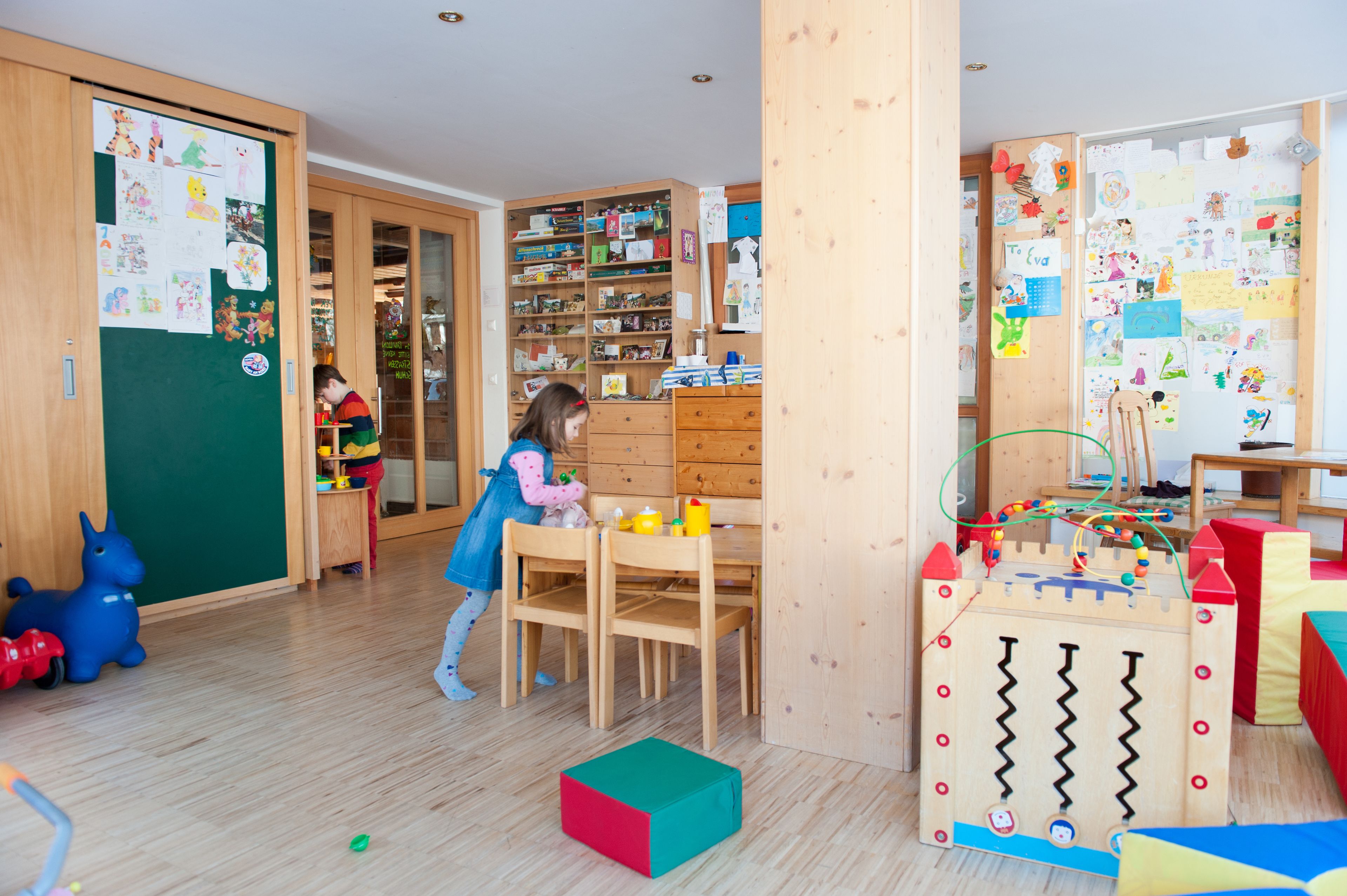 A room with wooden furniture, a bookshelf, and a bulletin board with children's drawings.