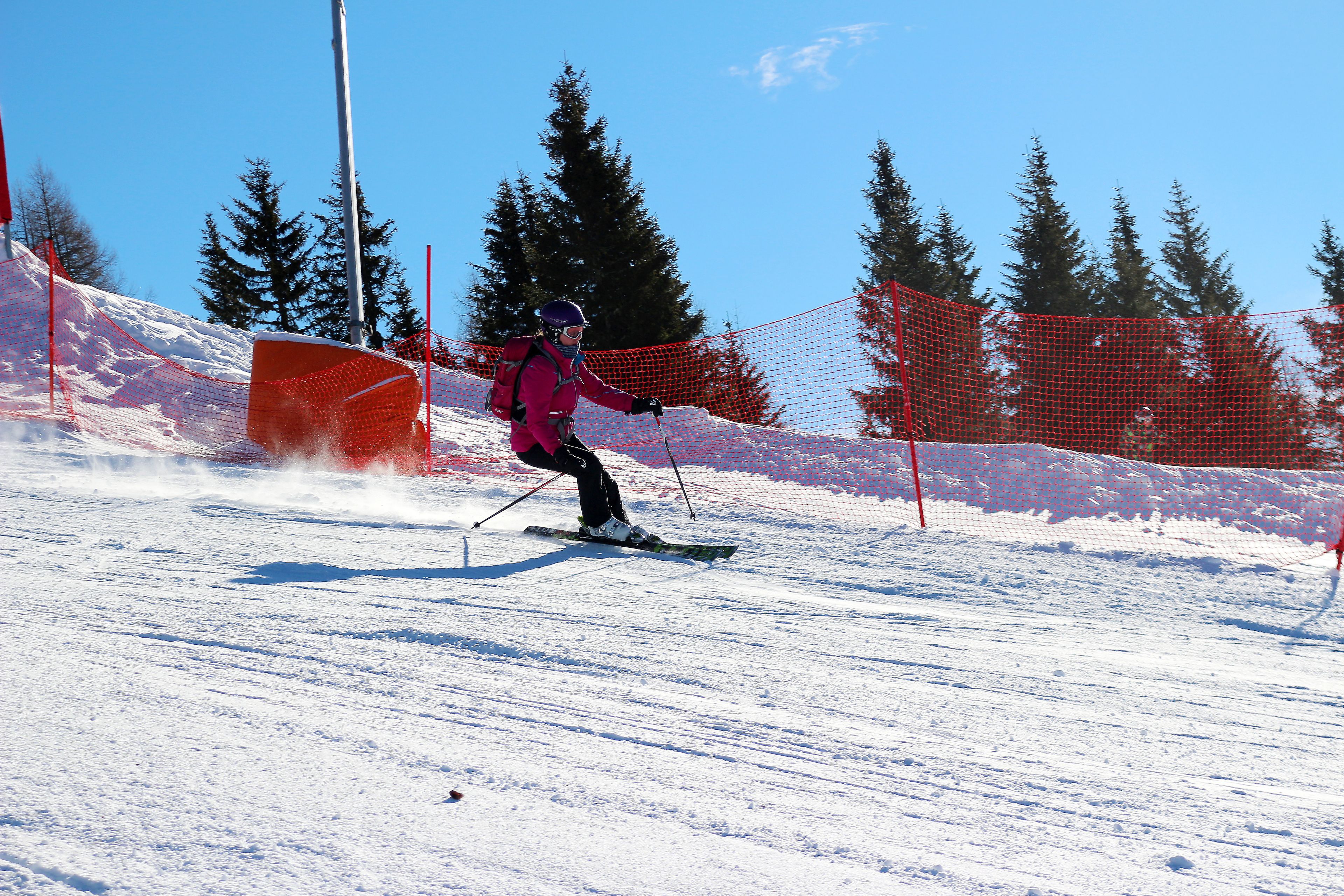 A person skiing on a snowy slope with orange safety nets and trees in the background.