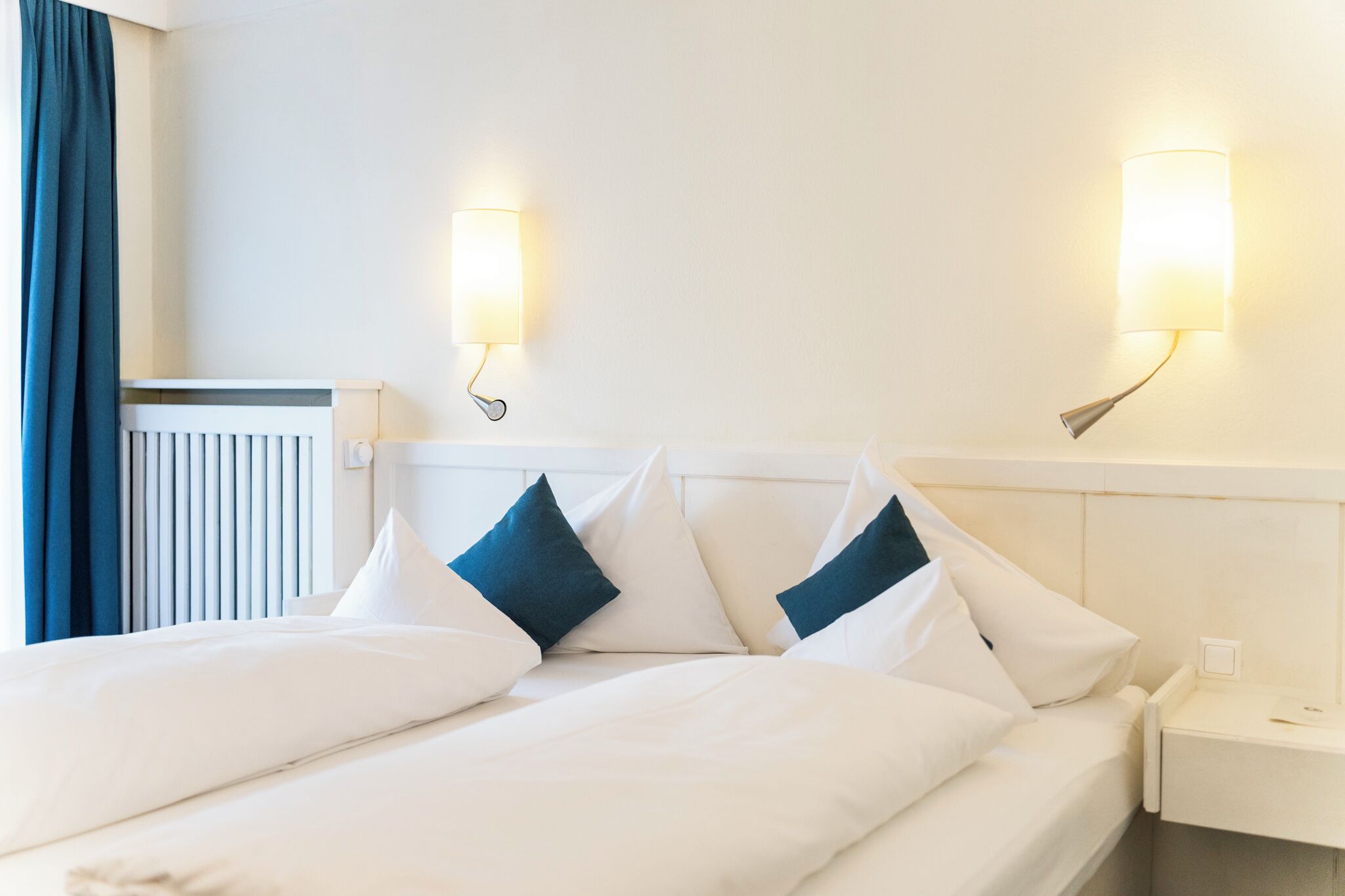 Ein Hotelzimmer mit einem ordentlich bezogenen Bett, zwei Wandleuchten und einem weißen Nachttisch.