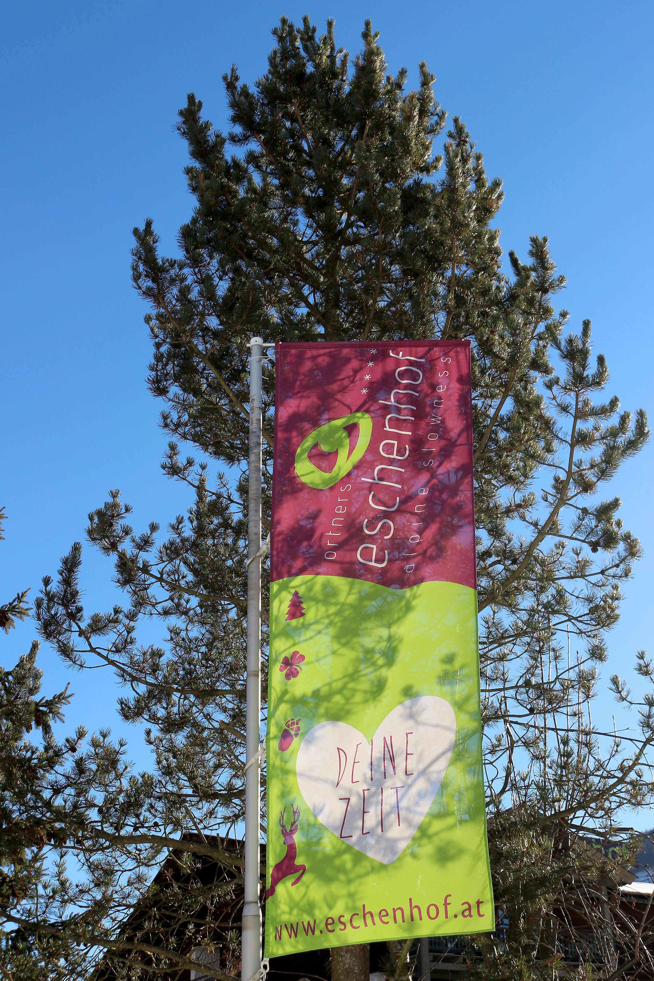 Een verticale banner met tekst en een website-URL, bevestigd aan een paal, met bomen op de achtergrond.