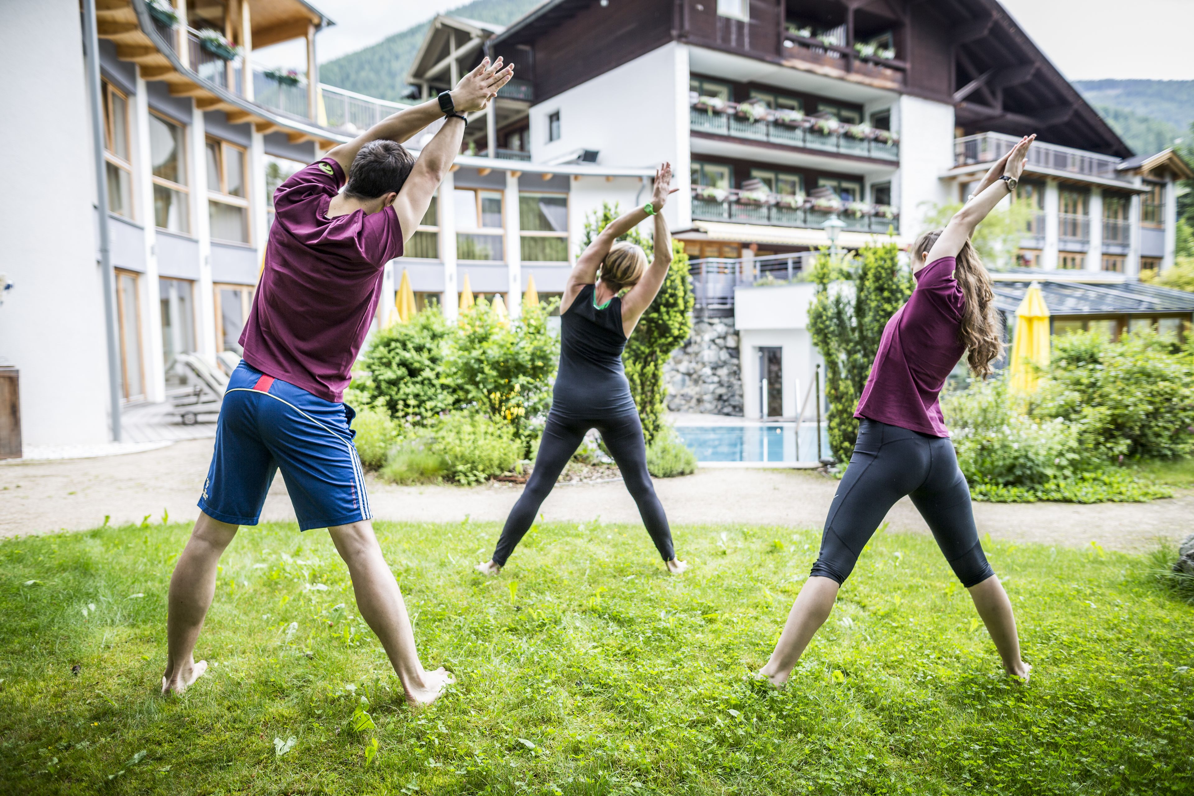 Three people performing a yoga pose outdoors in front of a building with balconies and a pool.