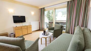 Hotelkamer met een bank, fauteuils, een kleine tafel met waterkoker, een televisie en uitzicht op een balkon met railing en bomen.