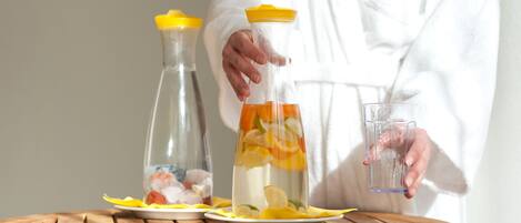 A person in a white robe pouring a drink from a clear bottle with a yellow cap into a glass on a wooden table.