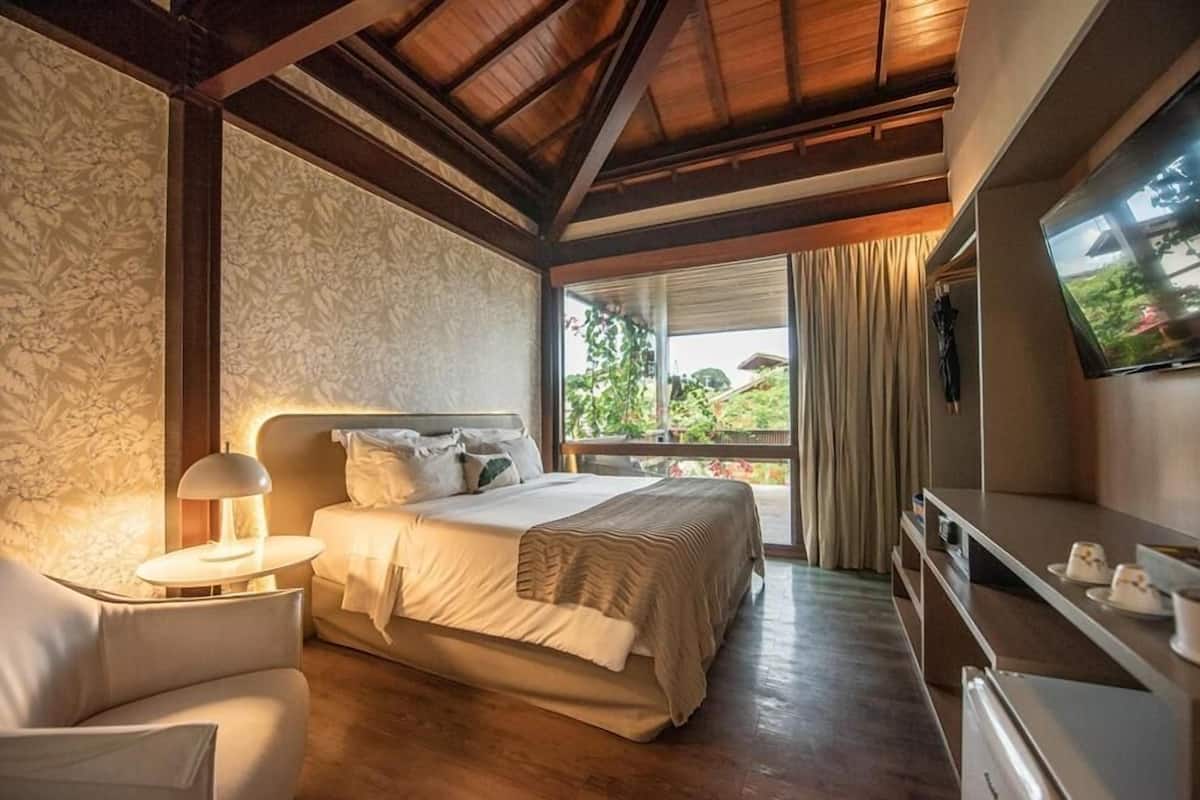 Un dormitorio con cama, mesita de noche, lámpara, televisor y vistas al verdor exterior.