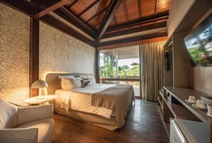 A bedroom with a bed, bedside table, lamp, TV, and a view of greenery outside.