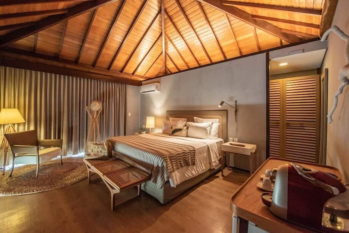 Un dormitorio acogedor con techo de madera, una cama grande, mesitas de noche, una silla y una pequeña mesa con lámpara.
