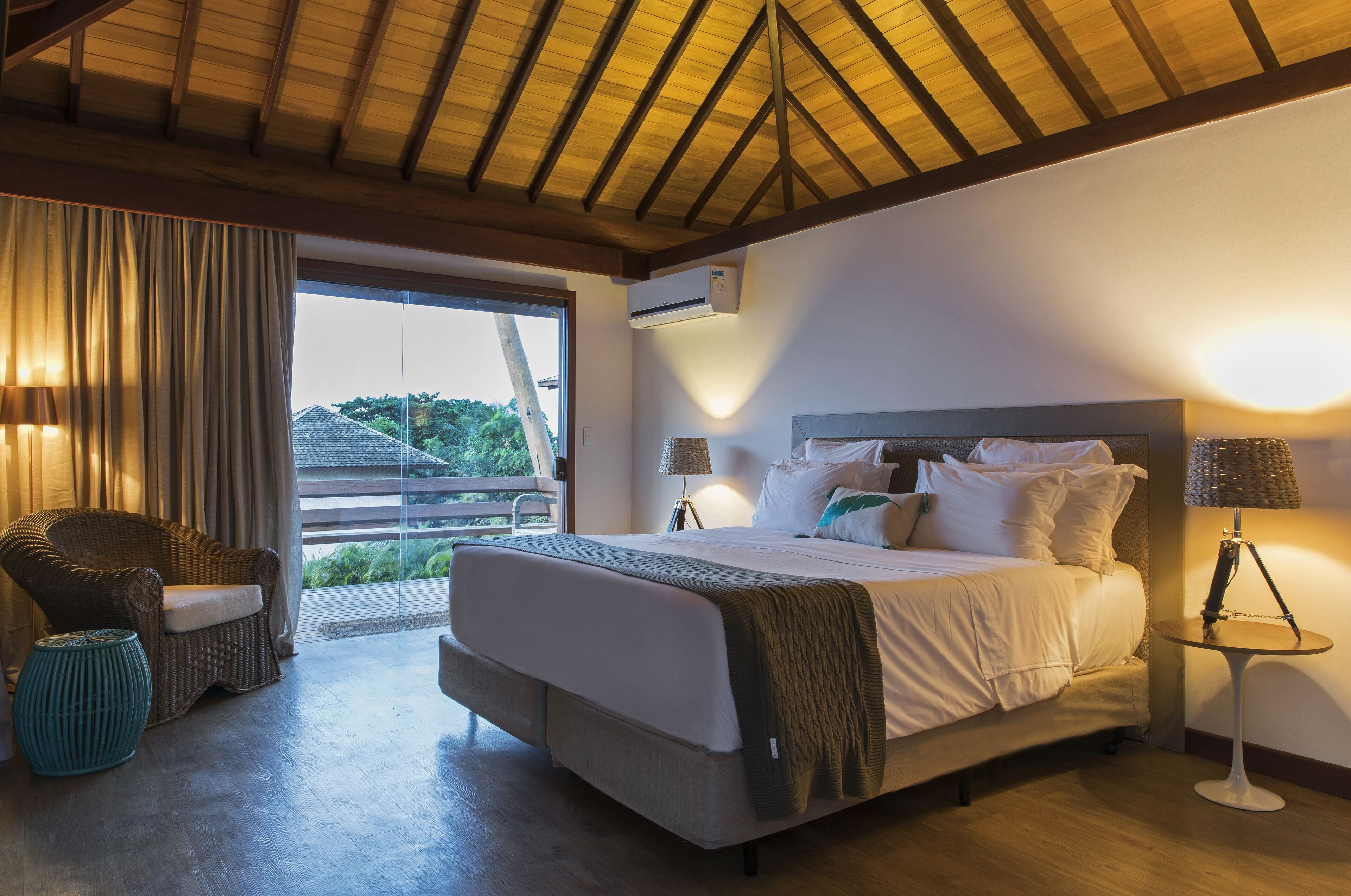 A bedroom with a large bed, a wicker chair, a small table, and a view of the outdoors.