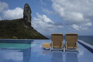 A poolside area with two wicker chairs and a small table, overlooking a body of water and a rocky island.